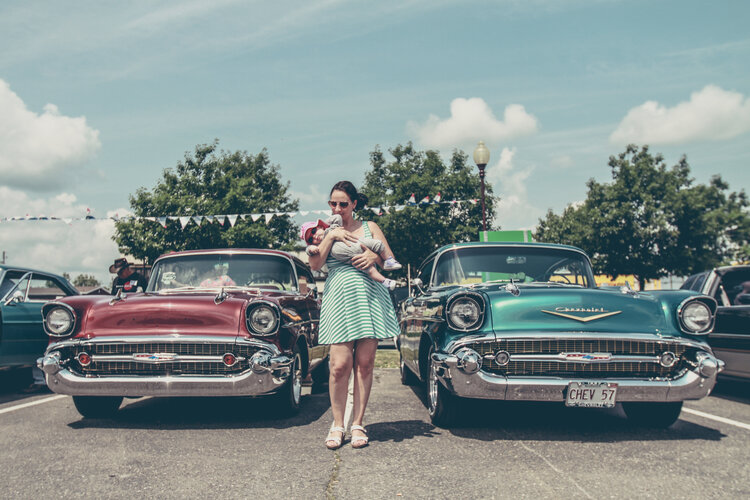 newborns. Mom in striped dress holding baby in front of 2 vintage cars
